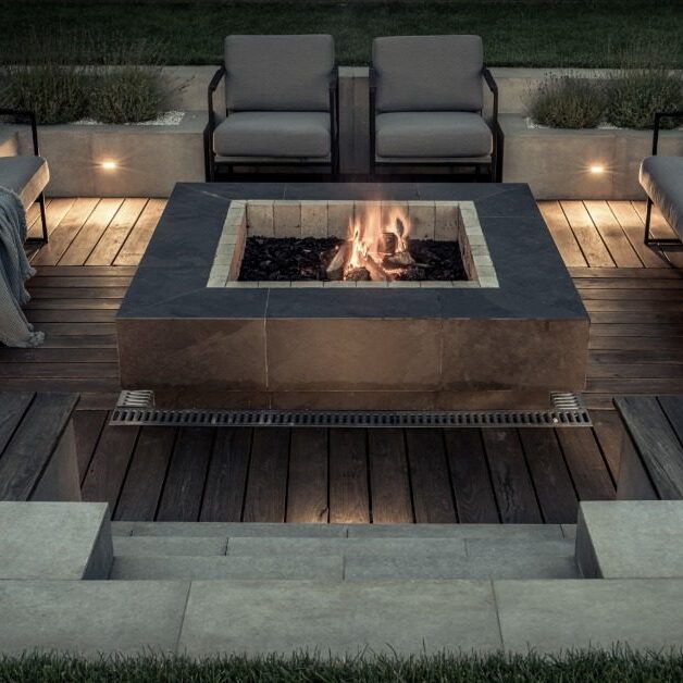 A rock square firepit in the middle of an outdoor area with couches and chairs, surrounded by grass and plants.