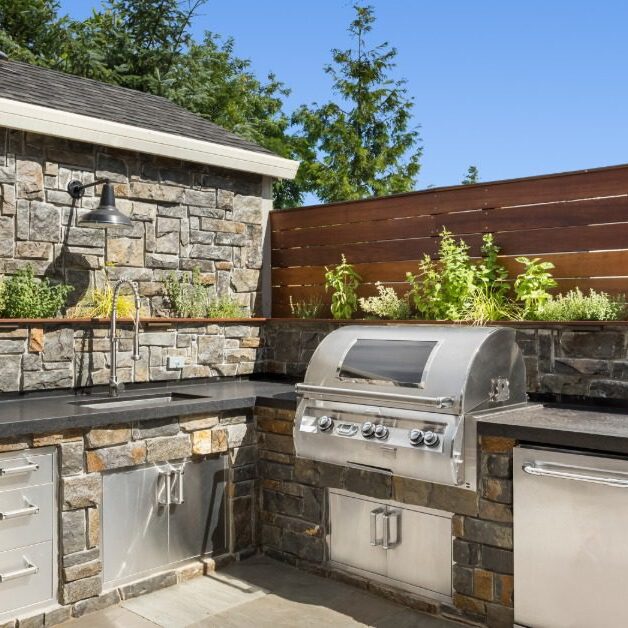 An outdoor kitchen made of rock, with a stainless-steel sink, a grill, a small refrigerator, and drawers on a sunny day.