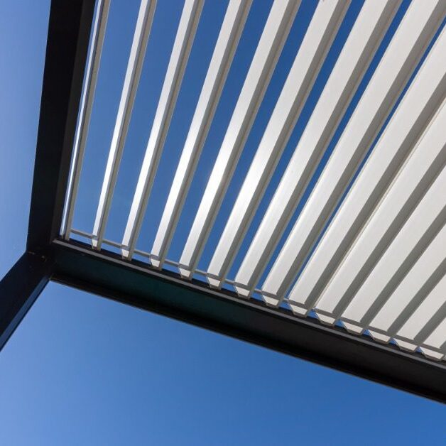 A ground view of an aluminum pergola with multiple thin white panels and thin legs, under the bright sun.
