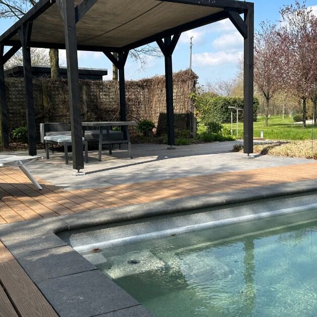 A pergola with six legs and a closed cover over a table and an outdoor couch, placed on a concrete base next to a pool.