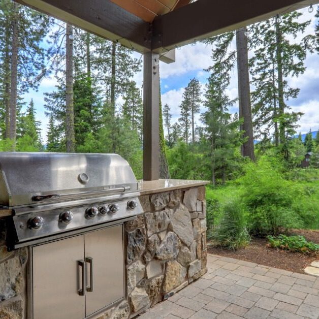 A stainless-steel grill with a large cover, built into a rock counter outdoors, under a pergola next to multiple trees.