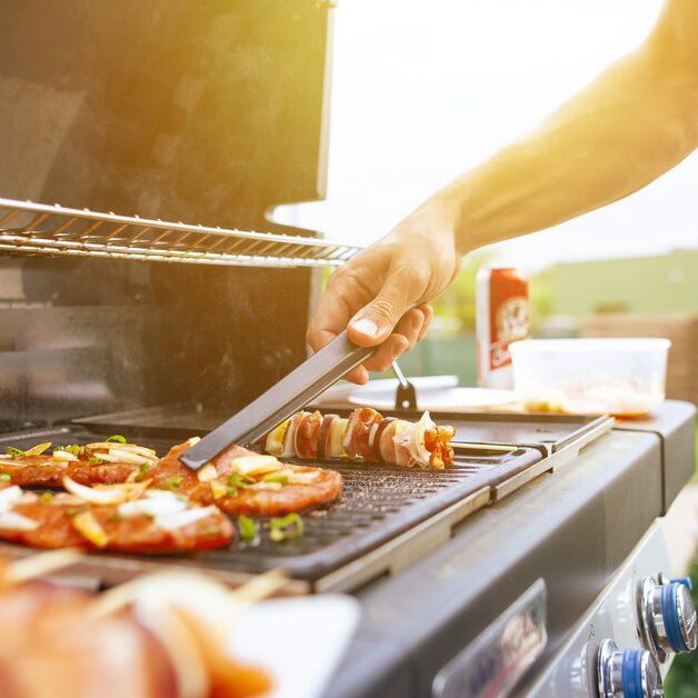 A close-up of the hands of a man using a pair of tongs to grab a piece of meat on a grill outdoors during a sunny day.