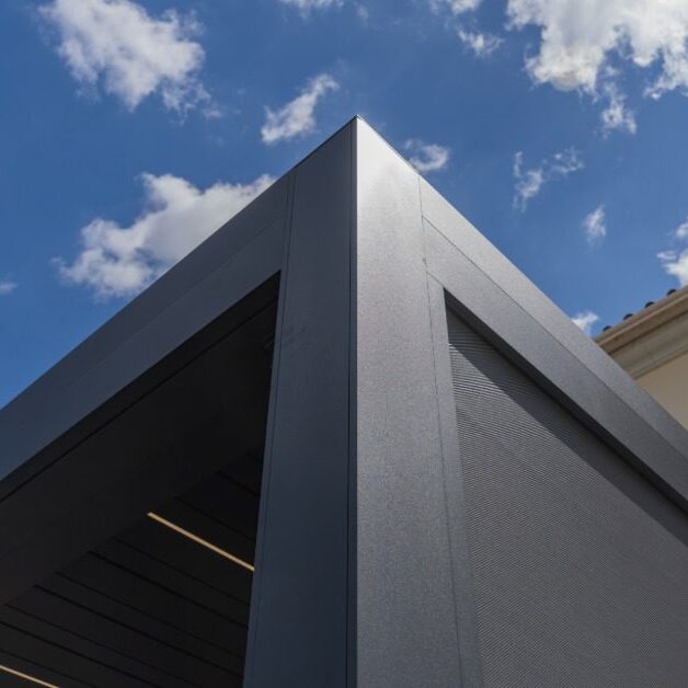 A close-up of the corner of a black aluminum pergola with multiple panels attached to the side of a two-story home.