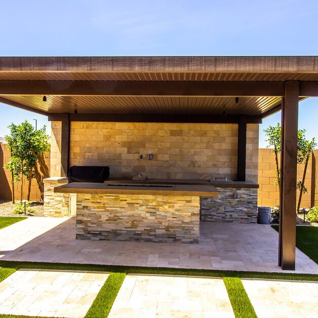 A wooden patio cover over an outdoor kitchen with an island and stainless steel appliances surrounded by grass.