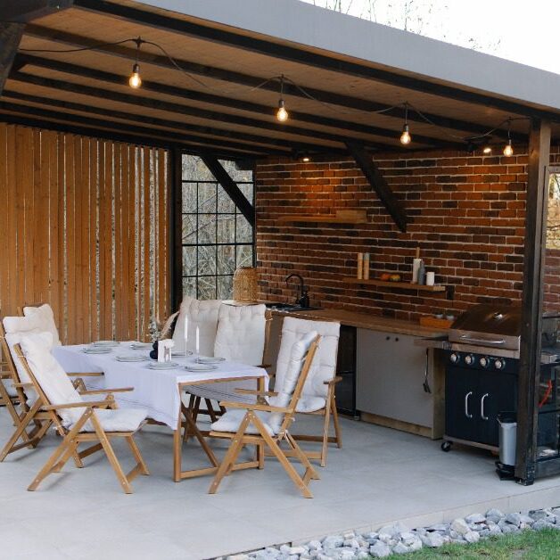 An outdoor kitchen next to a large dining table, under a pergola with hanging string lights over a concrete platform.