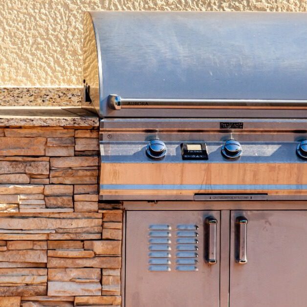 A stainless-steel grill with a rounded cover, knobs, and a storage compartment underneath, placed on a rock counter.