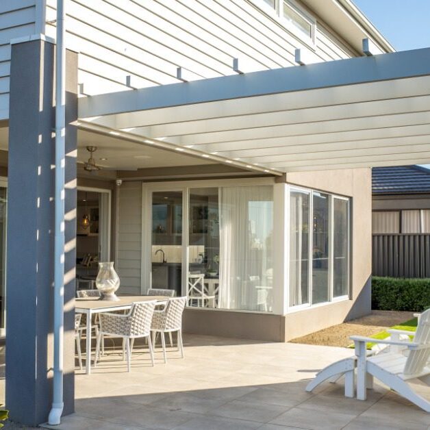 A two-story home with a large outdoor space under a white pergola with two lounging chairs and a dining table.