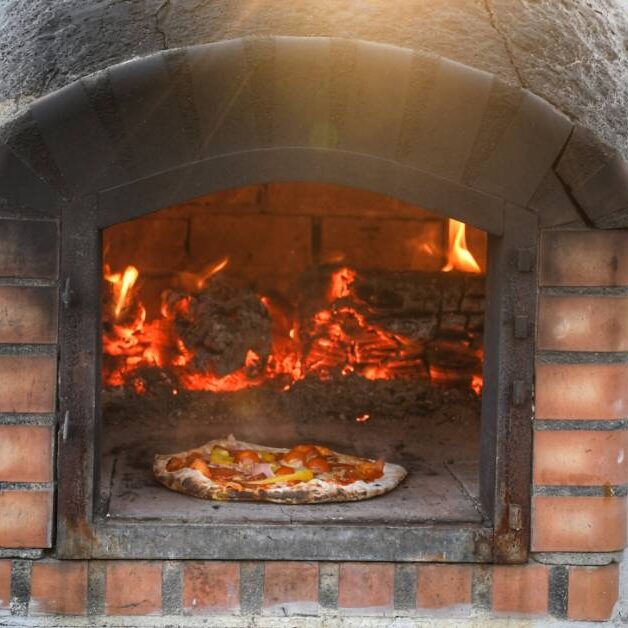 A close-up of a brick pizza oven with a rounded top and smoke marks, with burning wood inside, cooking a pizza outdoors.