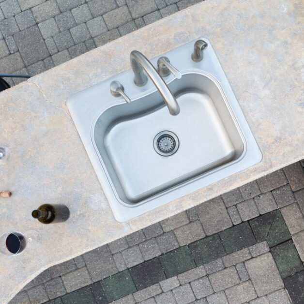 A white outdoor kitchen with black counters, a stainless-steel grill, and a sink, at the bottom of a white staircase.